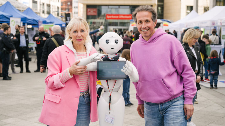 Ministerin Sinemus und Willi Weitzel ("Willi wills wissen") mit einer Figur eines Roboters, der ein Tablet in der Hand hält. 